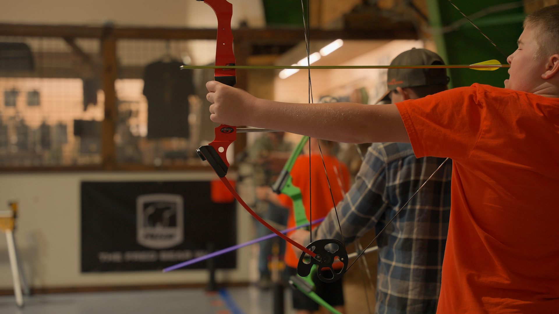 A boy in an orange shirt aiming a red bow and arrow indoors during archery practice.