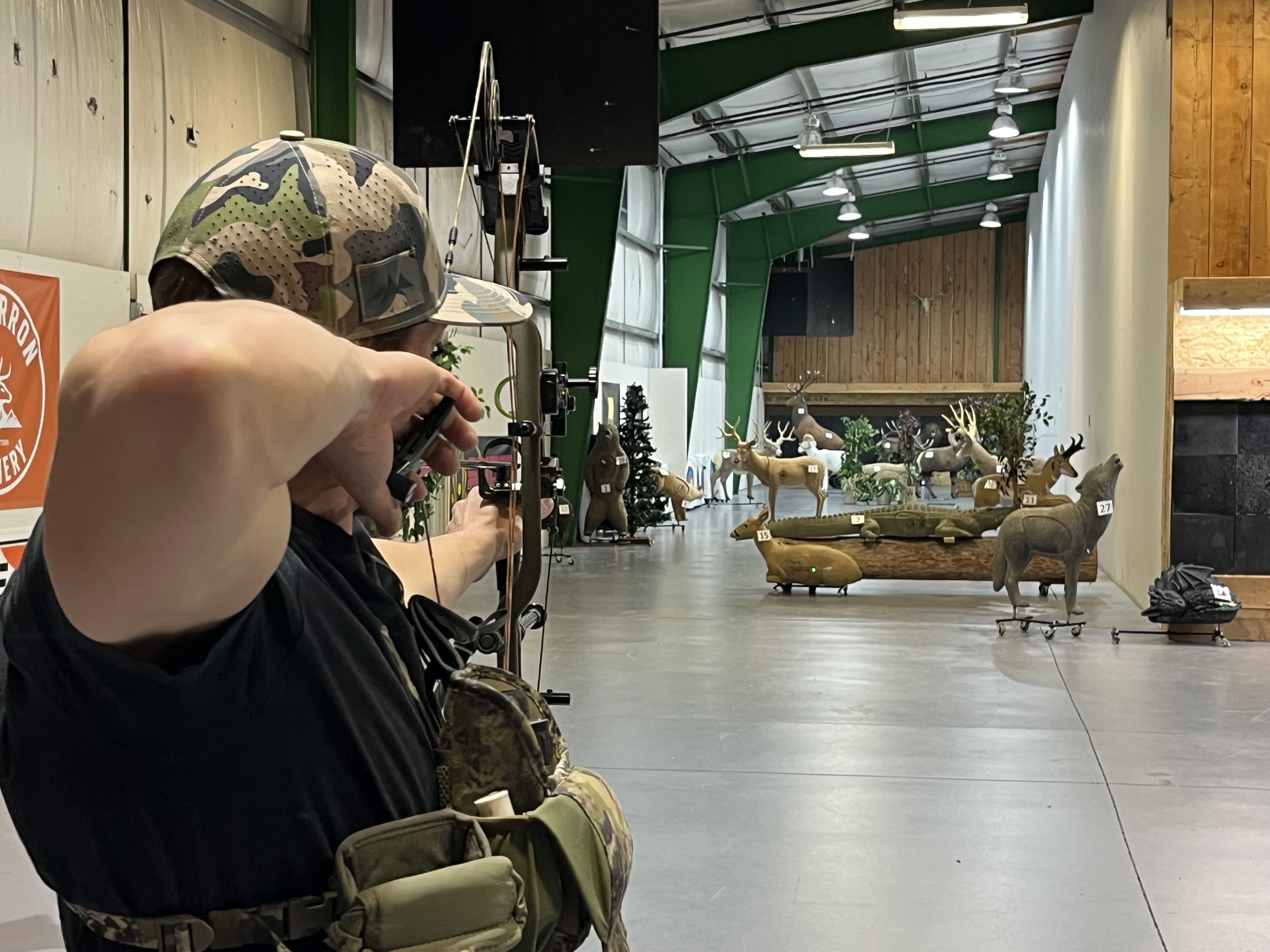 Person aiming a compound bow at animal-shaped targets in an indoor archery range.
