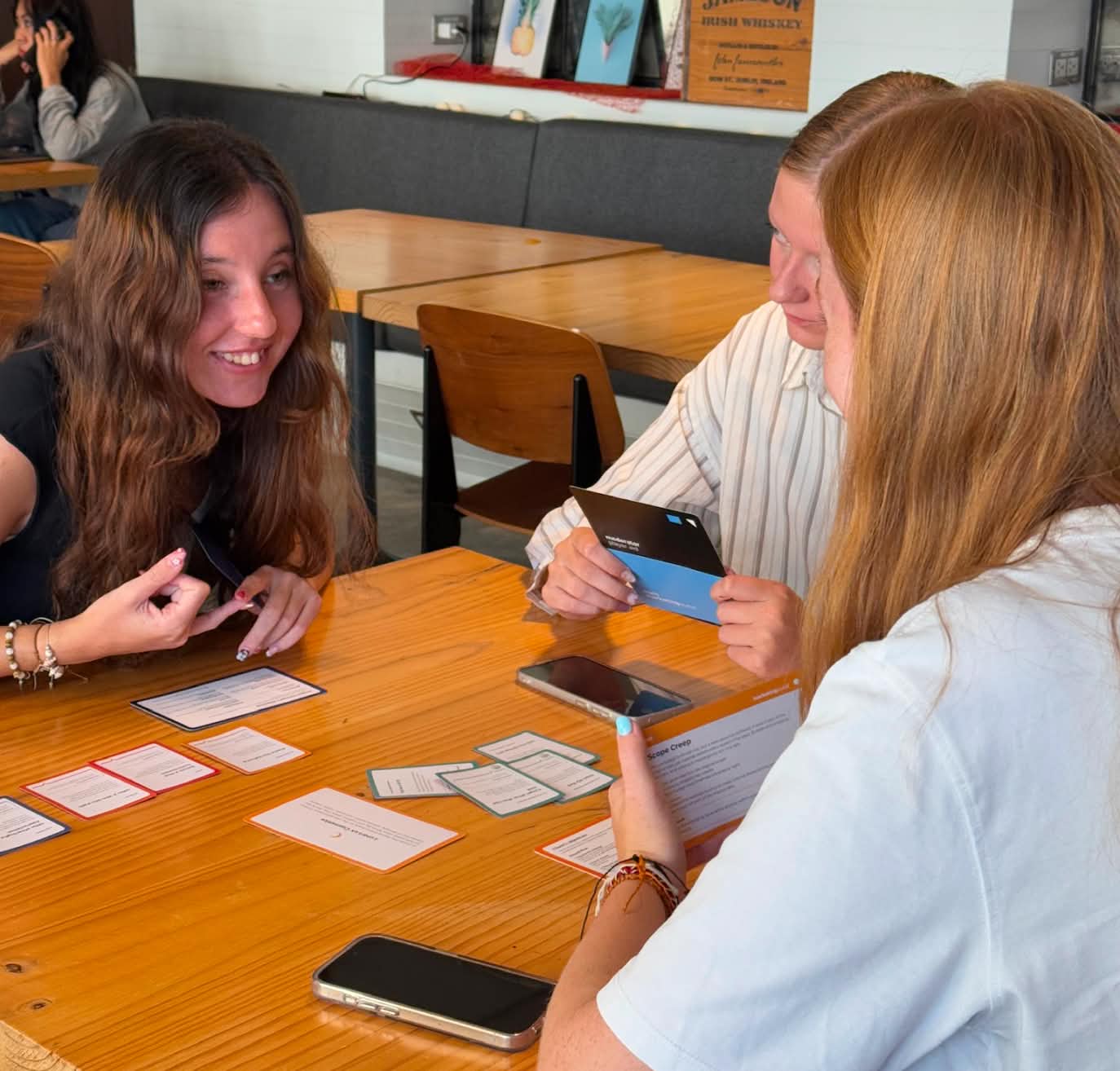 Three young women sitting at a wooden table playing a card game, smiling and engaged in conversation.