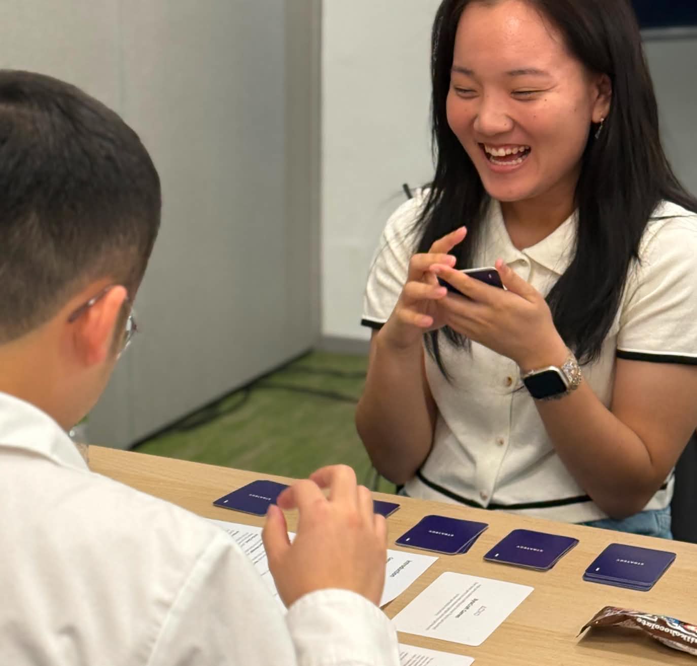 Two people sitting at a table playing a card game, with one woman smiling and holding cards while the man looks at cards laid out on the table.