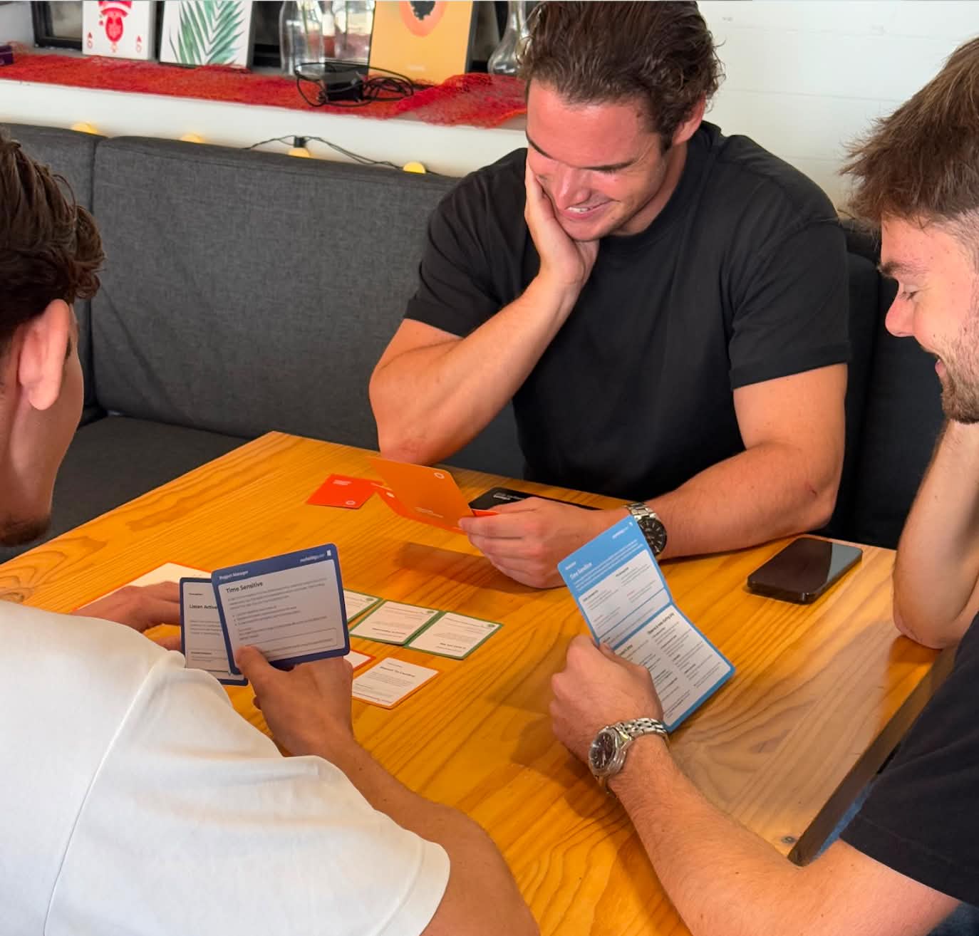 Three young men sitting around a wooden table playing a card game with cards and instruction sheets.