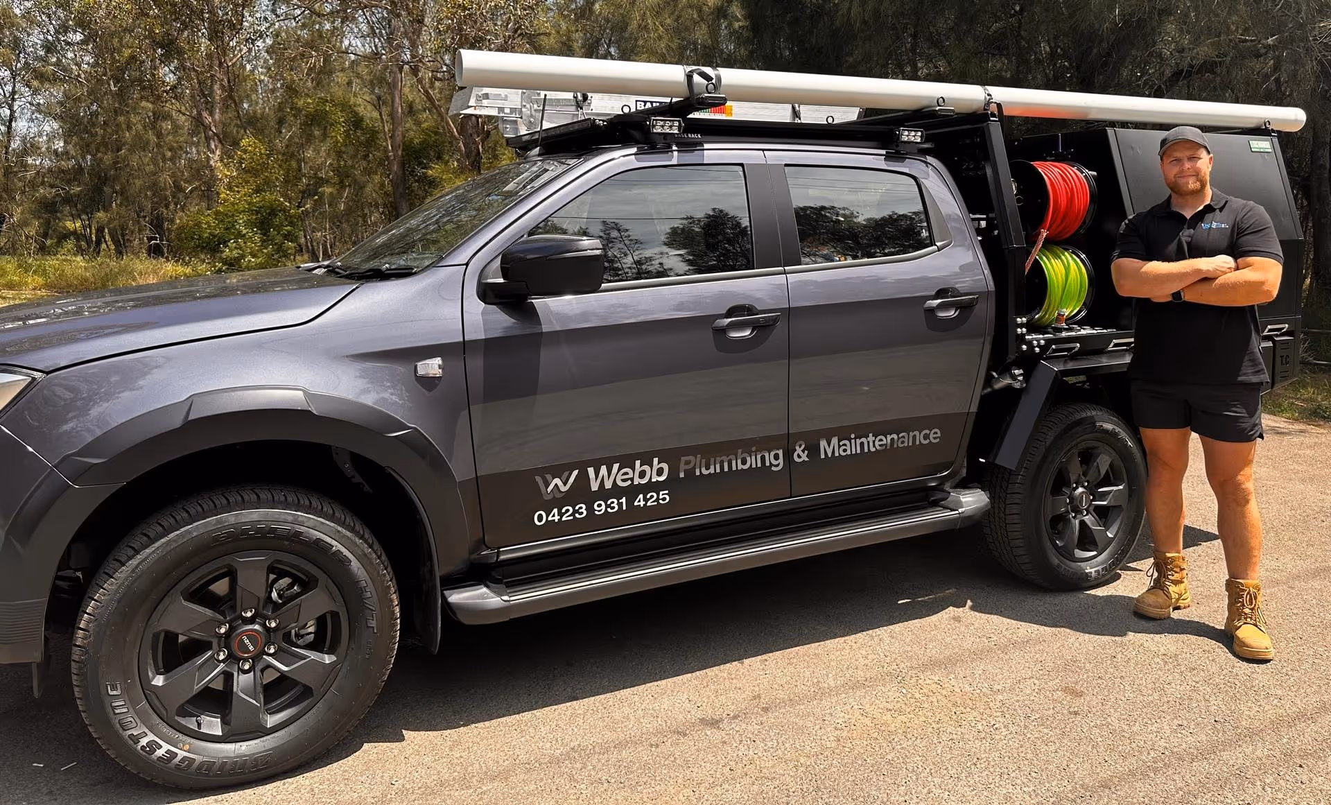 Man standing with arms crossed next to a gray pickup truck labeled Webb Plumbing & Maintenance, equipped with plumbing hoses and tools.