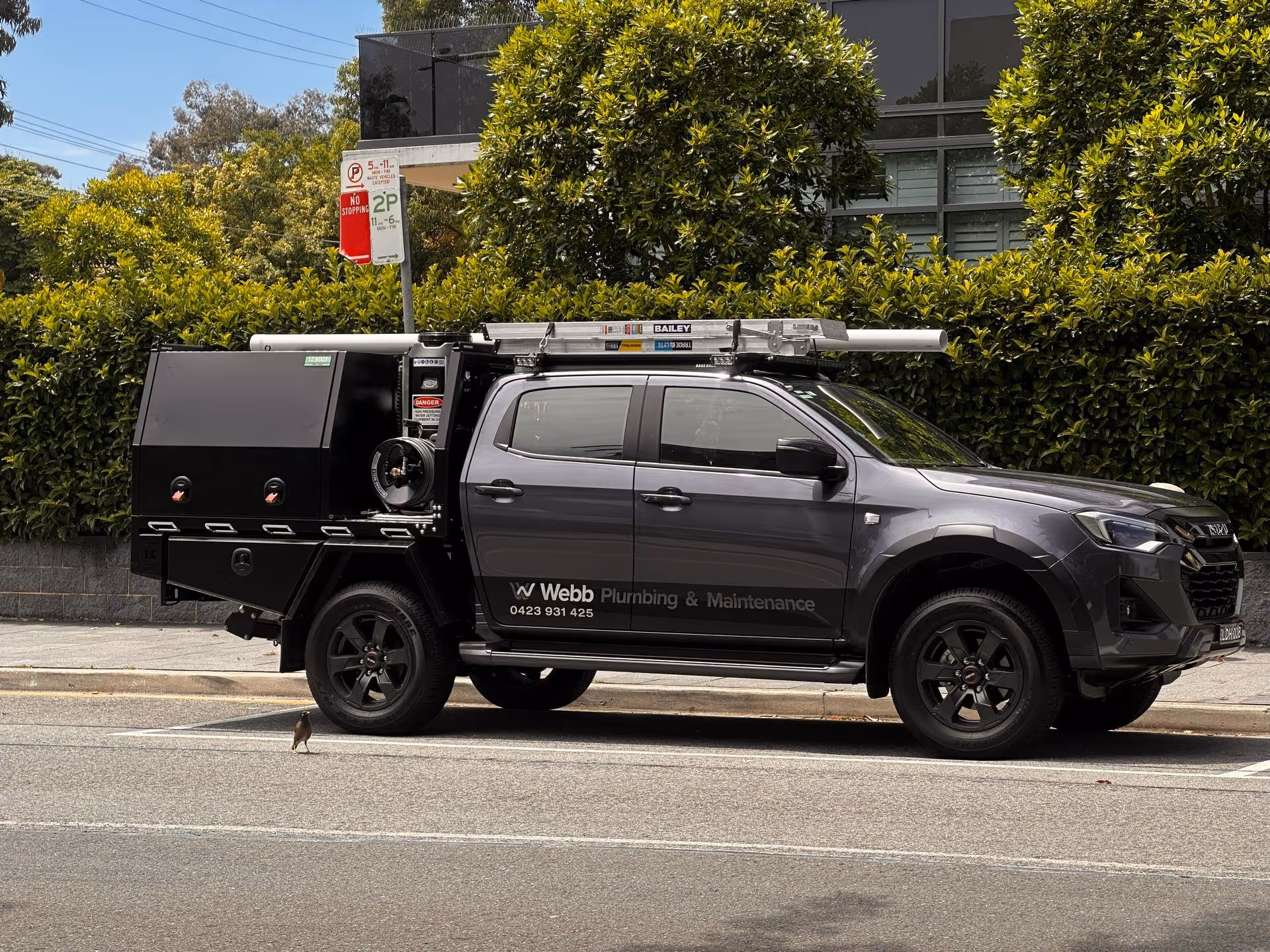 Gray pickup truck with plumbing maintenance equipment and logo parked on street in front of green bushes.