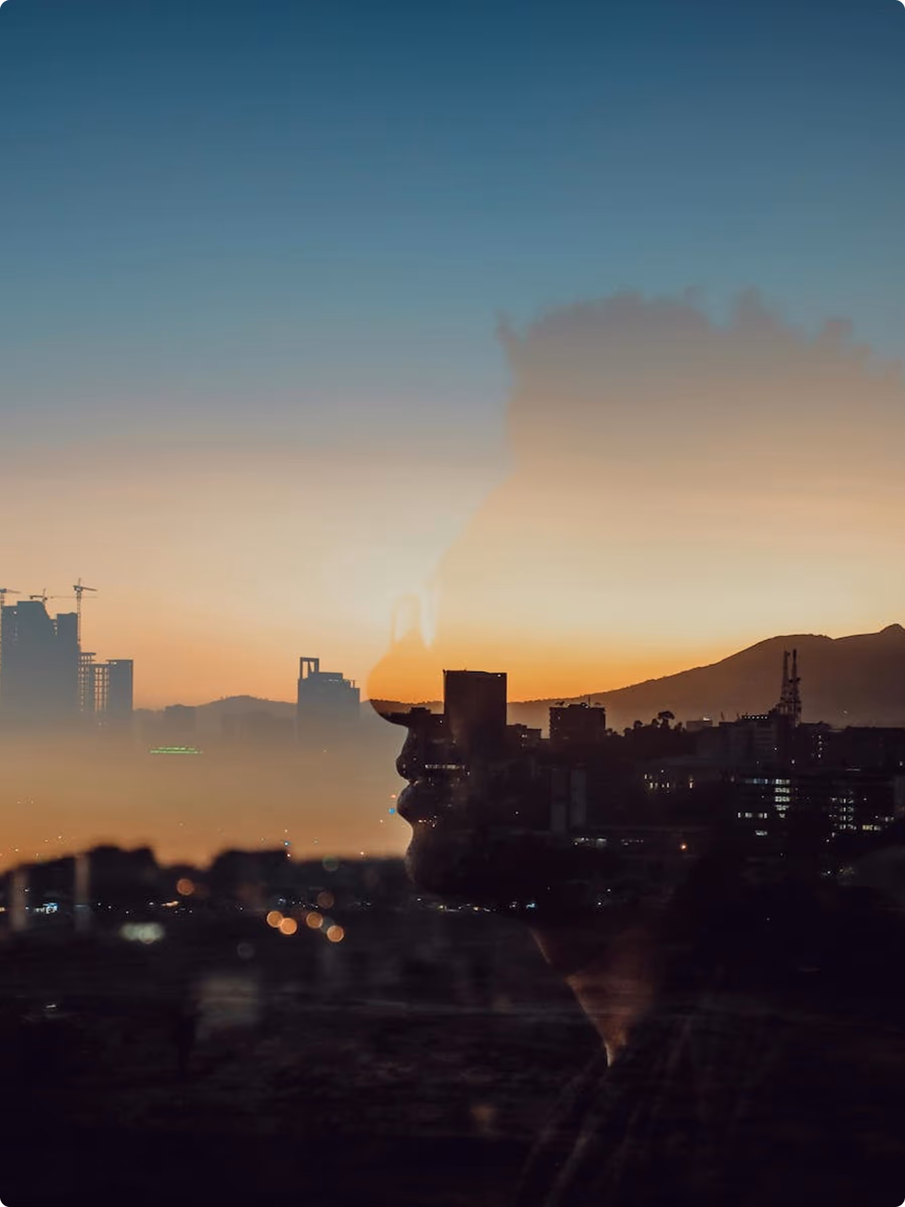 Double exposure profile of a man wearing a hat over a city skyline at sunset with mountains in the background.