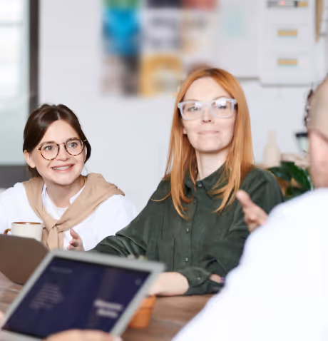 Two women wearing glasses and smiling while sitting at a table during a meeting, with a person holding a laptop in the foreground.