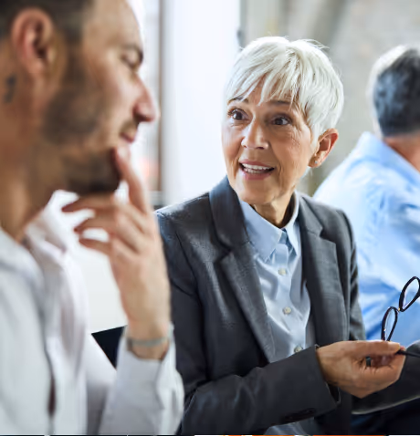 Two women wearing glasses and smiling while sitting at a table during a meeting, with a person holding a laptop in the foreground.