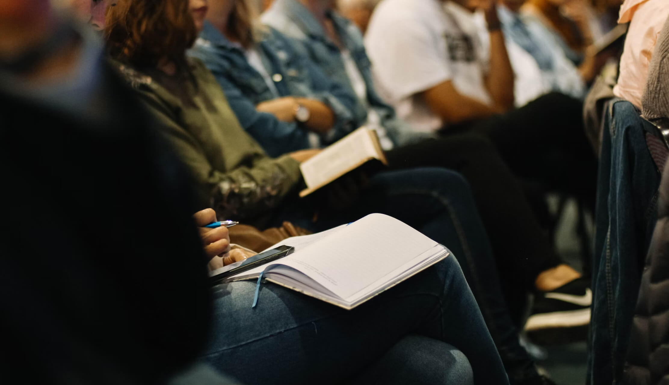 People seated closely together, some taking notes with notebooks and pens, others reading books during a session.