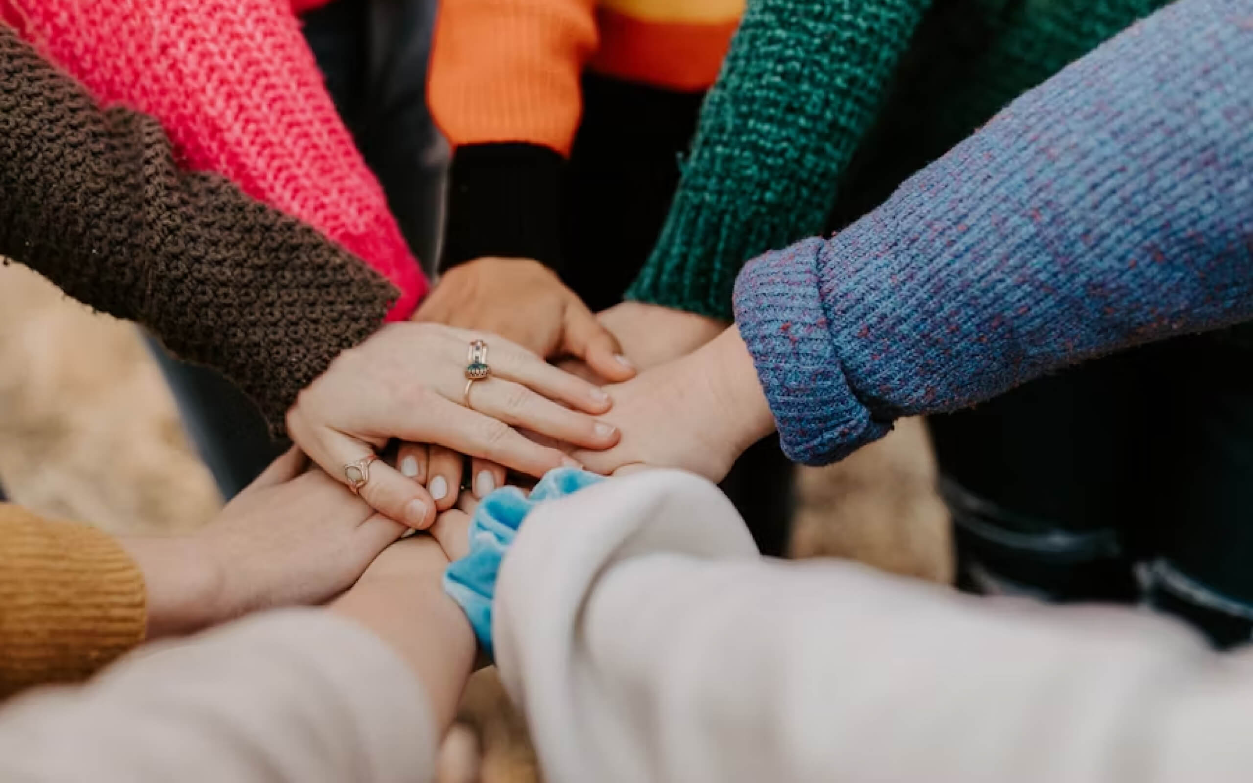 Multiple hands stacked together in a teamwork gesture with diverse sweater colors visible.