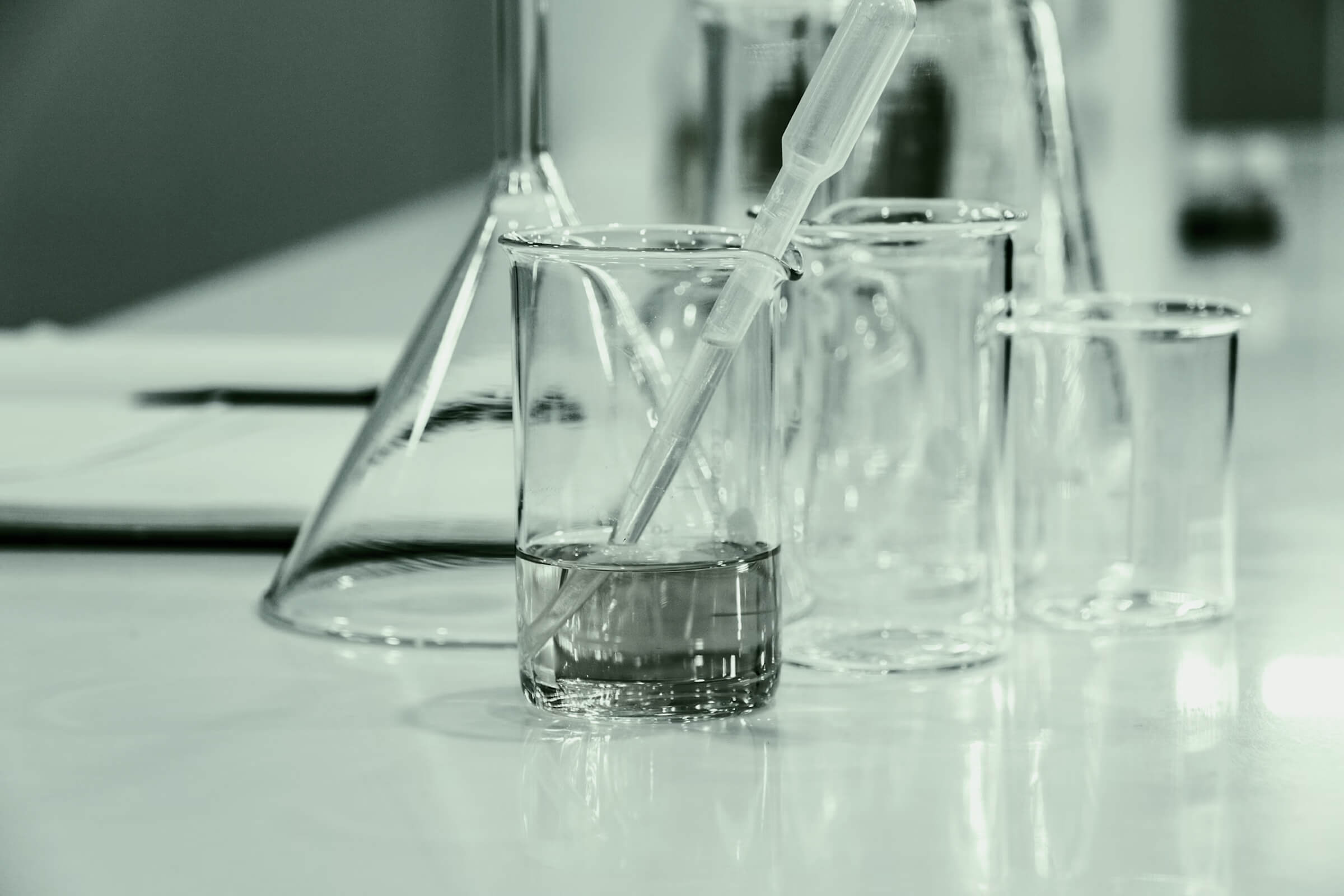 Close-up of laboratory glassware including a beaker with liquid and a pipette, alongside other empty beakers and flasks on a lab bench.