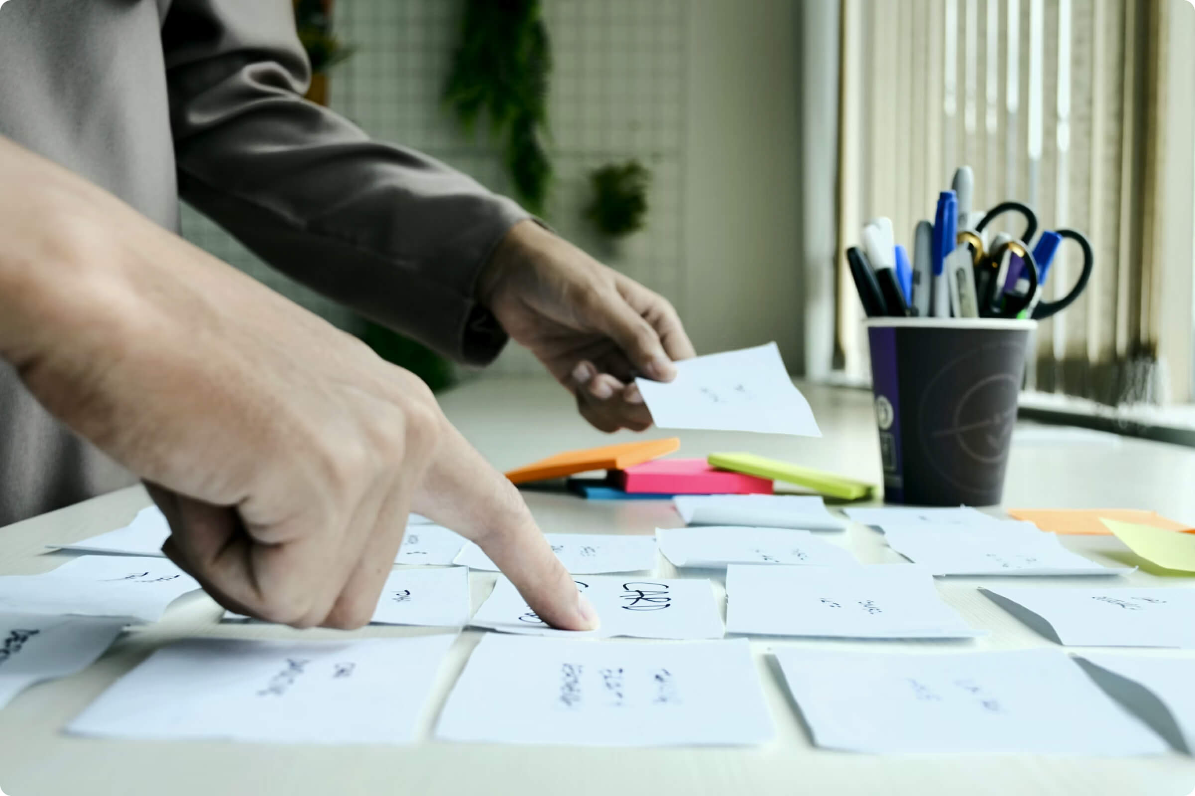 Person organizing handwritten notes on a table with sticky notes and a cup of pens and scissors nearby.
