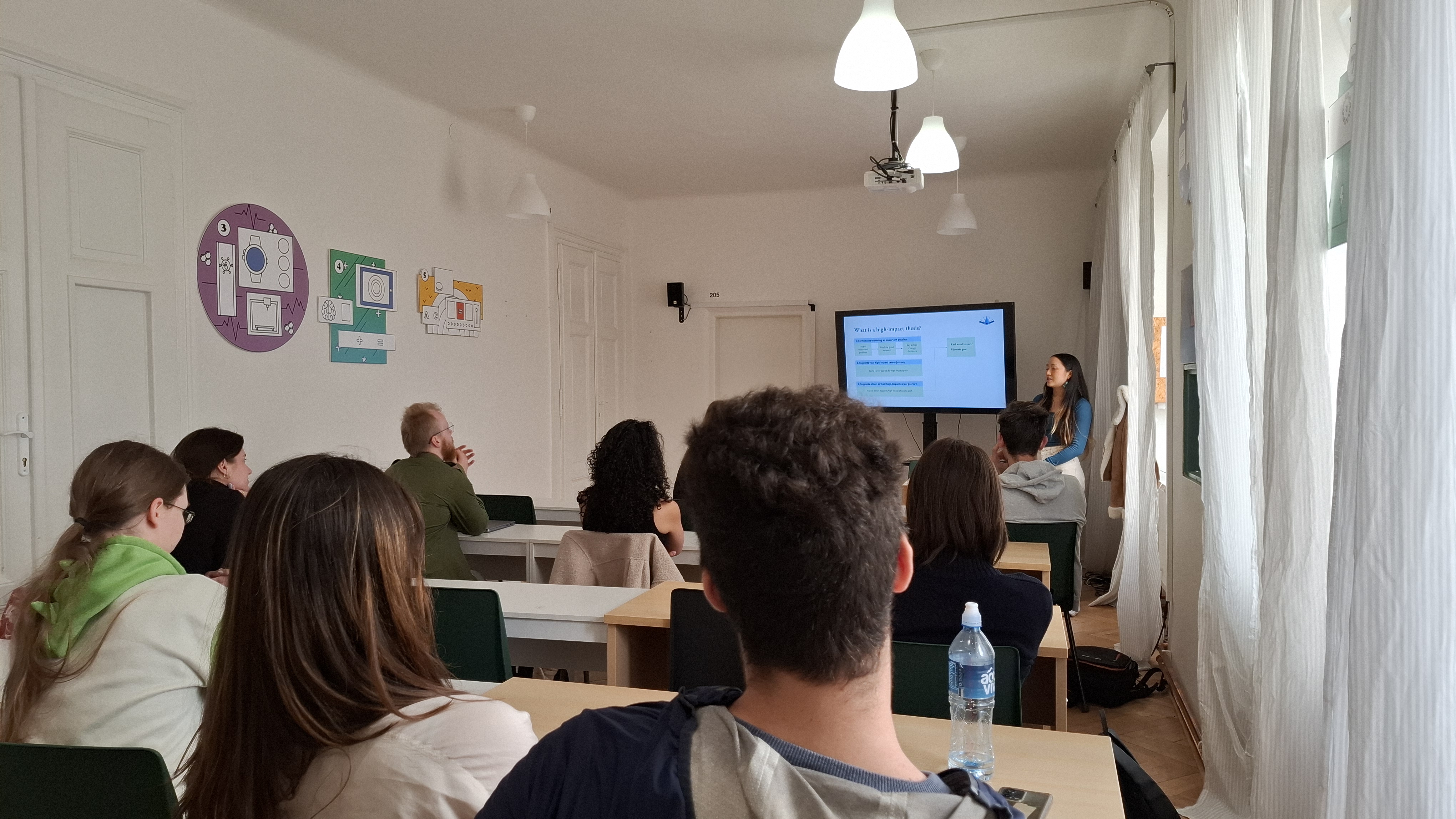 Classroom with students seated facing a teacher who is presenting a slide about a high-impact thesis on a screen.
