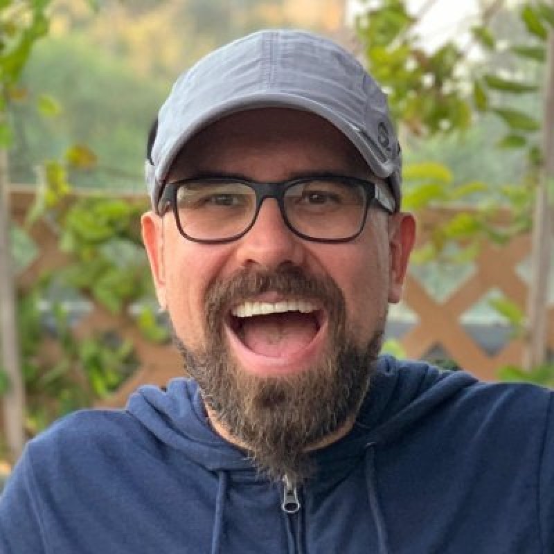 Smiling man with a beard, glasses, and a light gray cap wearing a navy blue hoodie outdoors with greenery background.