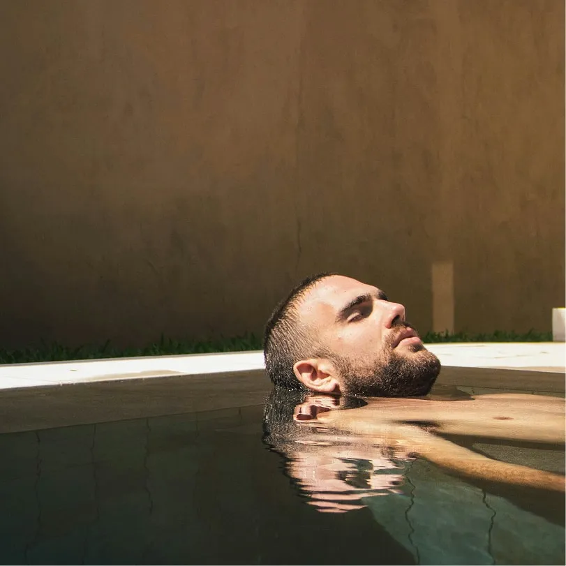 Man with a beard relaxing in a pool, partly submerged in water with his head tilted back.