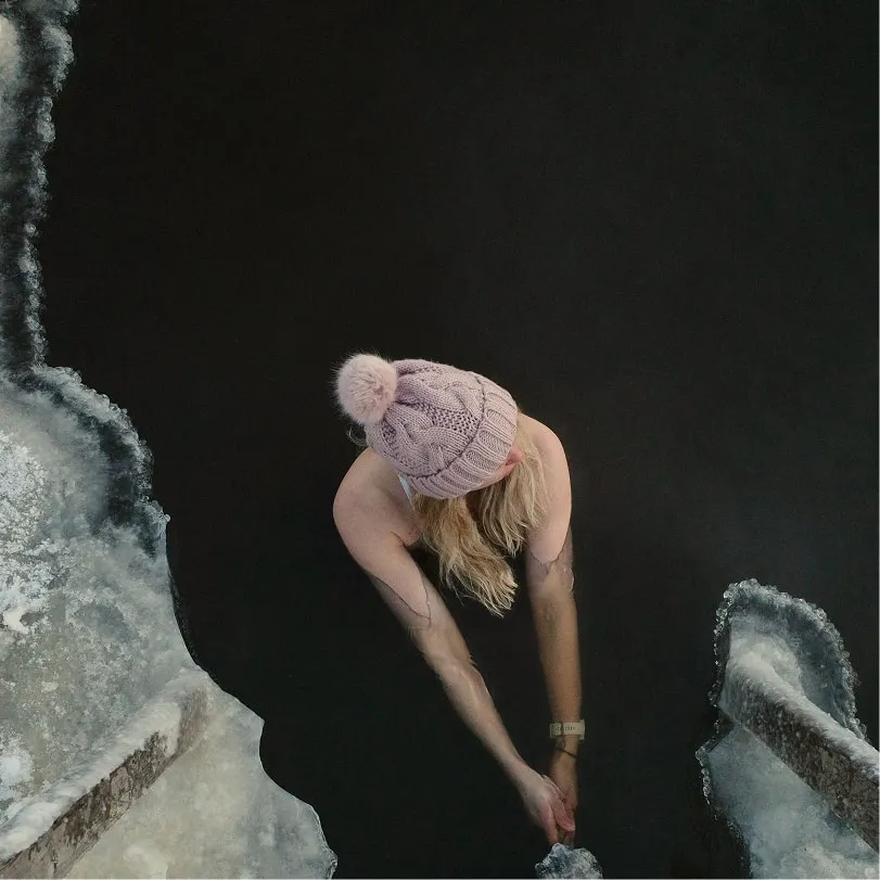 Top-down view of a person wearing a pink knit hat with a pom-pom, reaching their hands into dark water surrounded by ice.