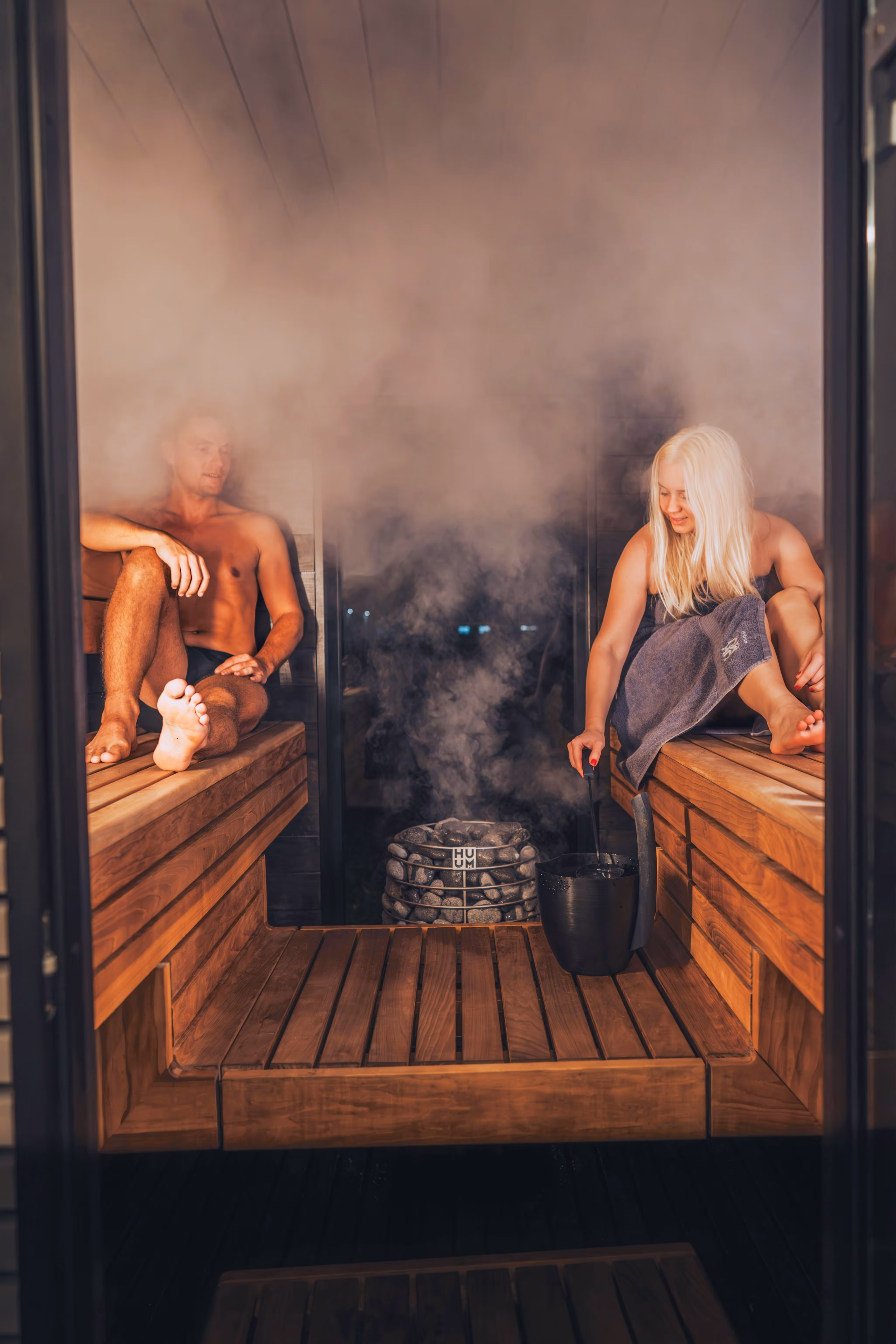 A shirtless man and a blonde woman wrapped in a towel sitting on wooden benches inside a steam-filled sauna, with the woman pouring water onto hot stones.