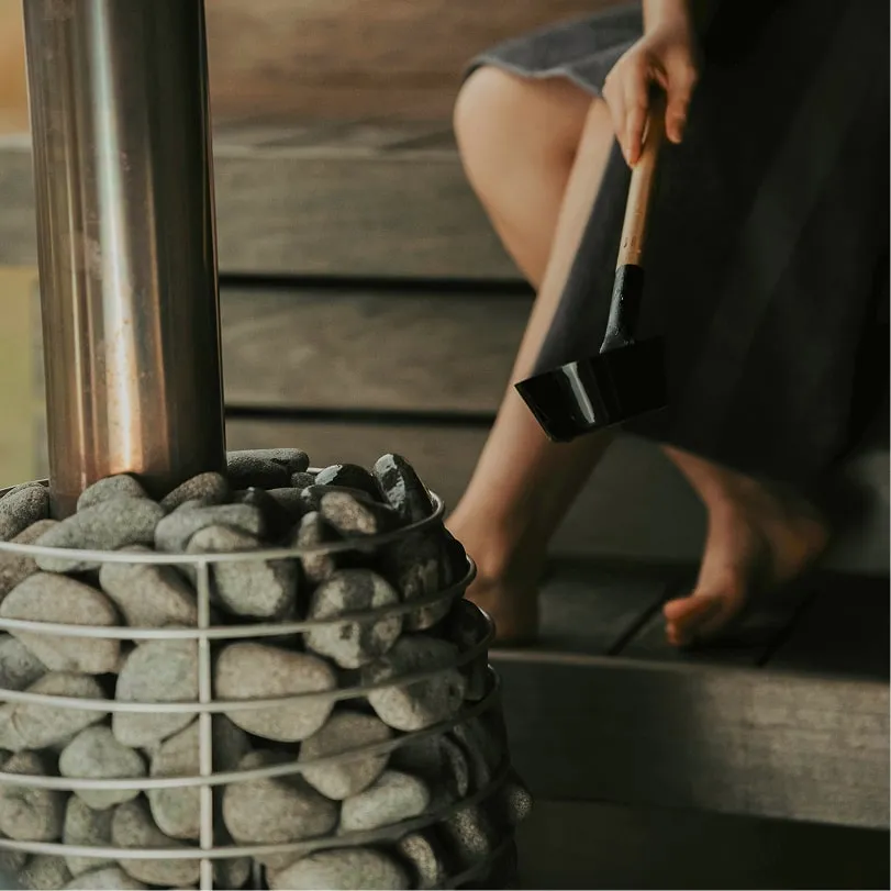 Person sitting barefoot near a stone-filled sauna heater holding a wooden ladle.
