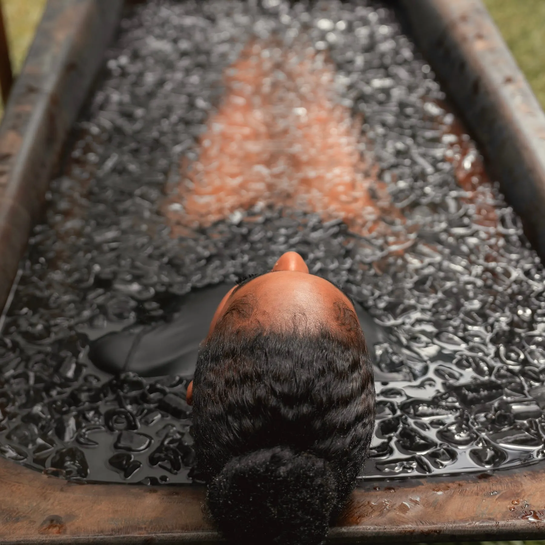 Person with tightly coiled hair tied back, submerged up to their neck in a wooden tub filled with ice cubes.