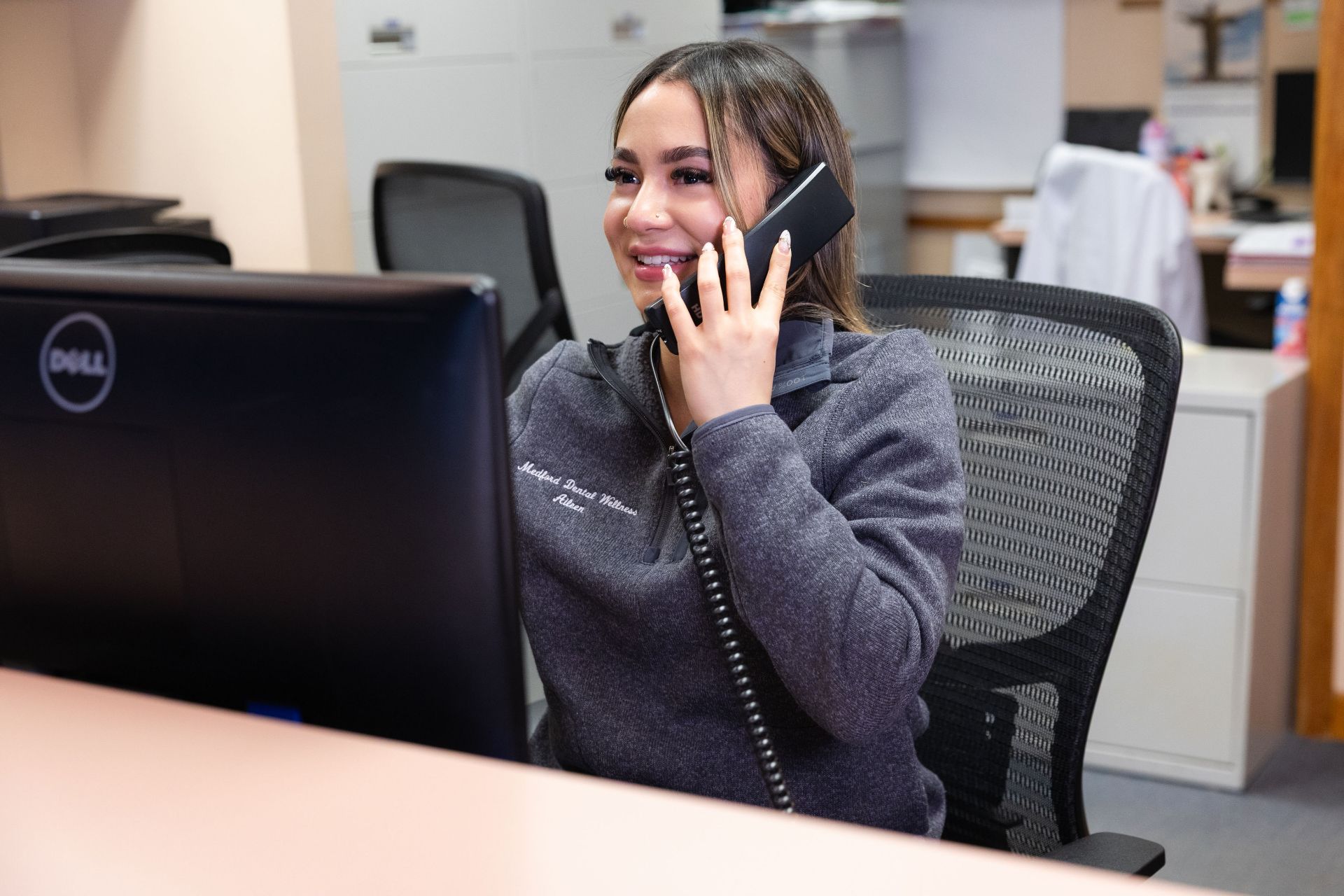 Smiling woman speaking on a corded phone at her office desk in front of a computer.