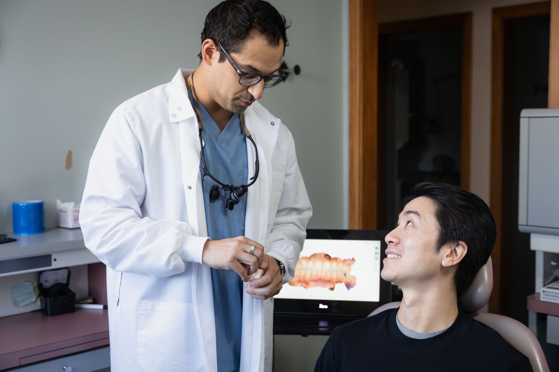 Dentist in white coat preparing dental tools while talking to a smiling patient sitting in the dental chair.