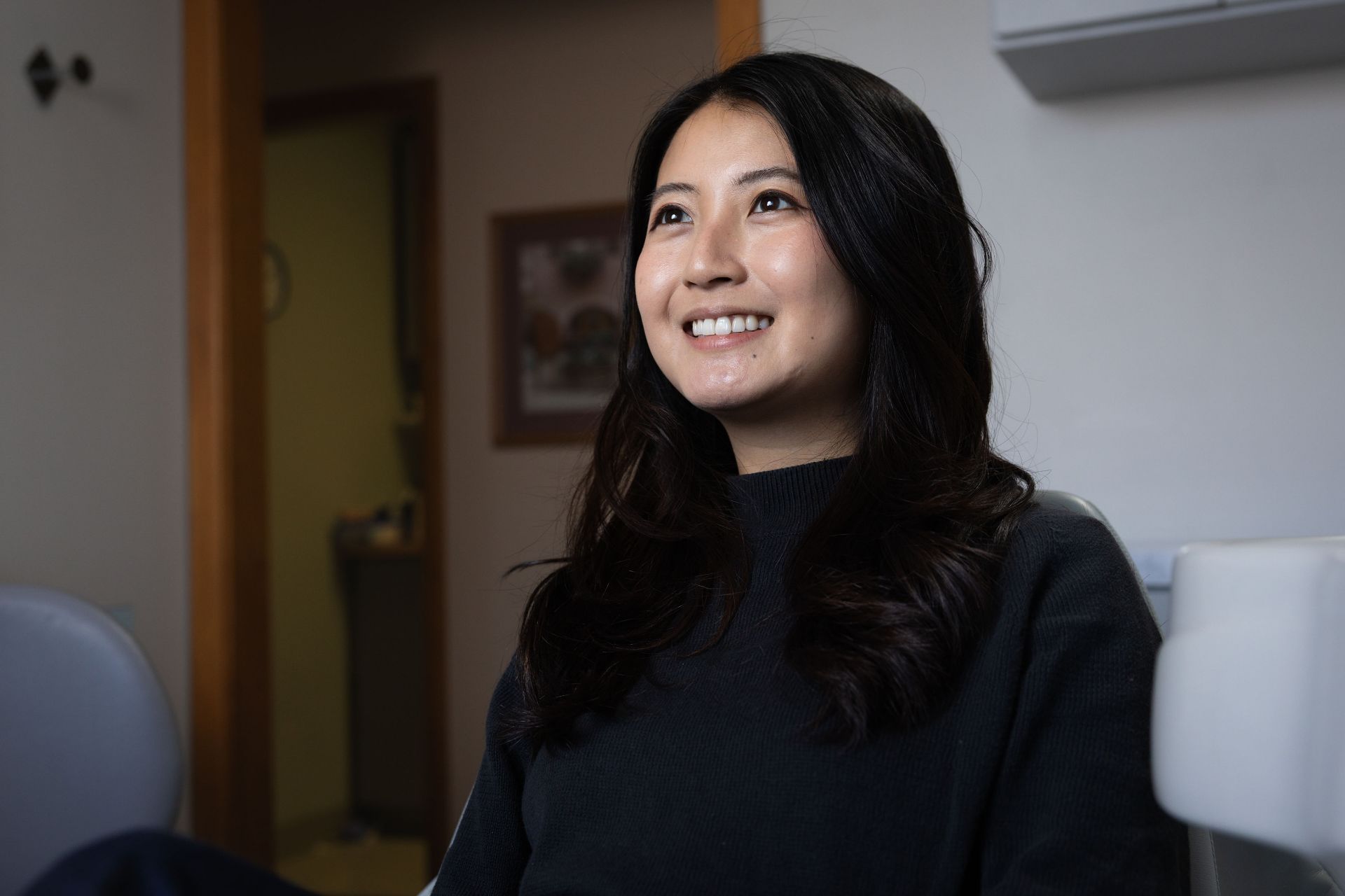 Smiling woman with long black hair wearing a black sweater sitting indoors.