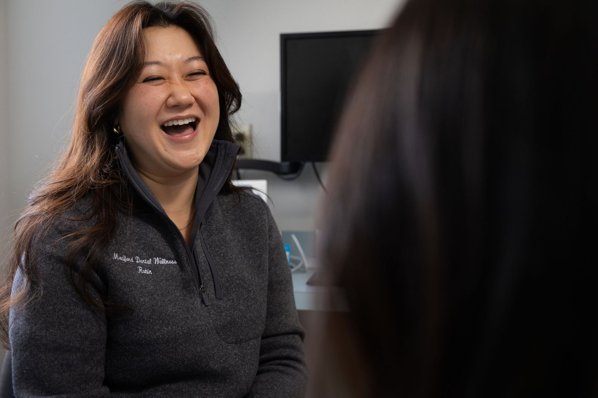 A woman wearing a dark zip-up jacket embroidered with 'Medford Dental Wellness Robin' smiling and laughing indoors.