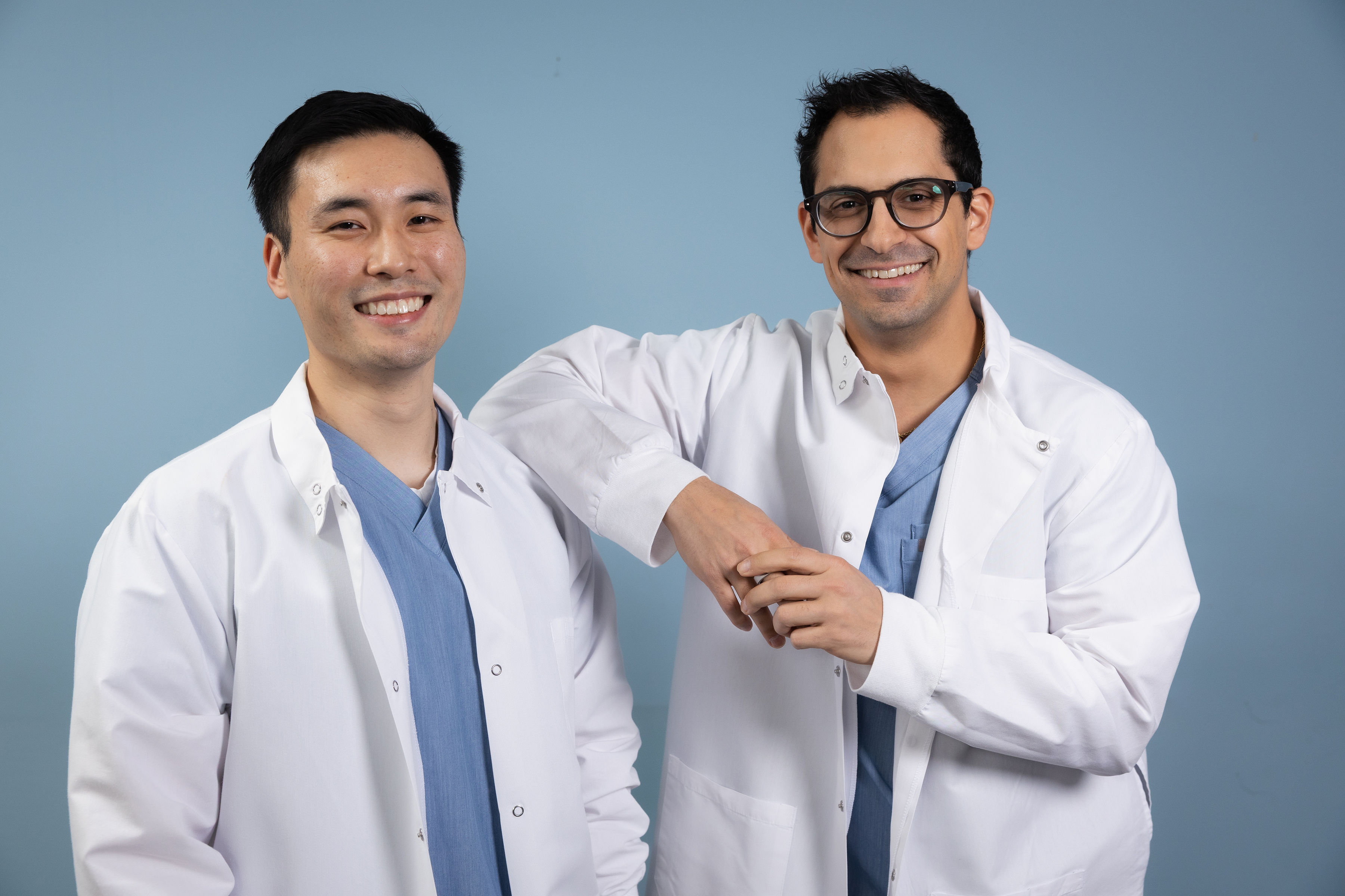 Two male medical professionals in white lab coats and scrubs smiling against a light blue background.