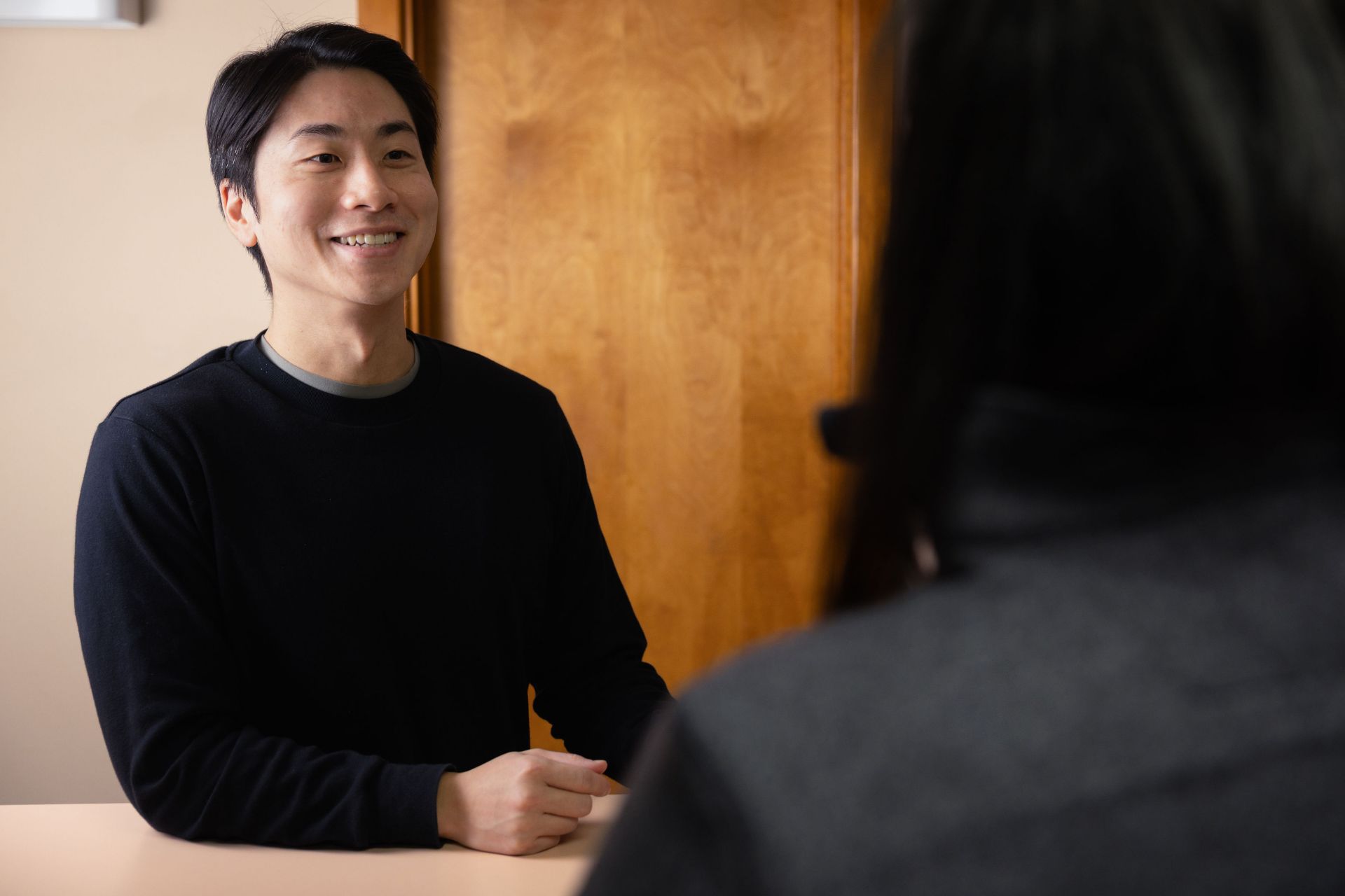 Smiling man in black sweater sitting at a table talking to a person with dark hair in the foreground.