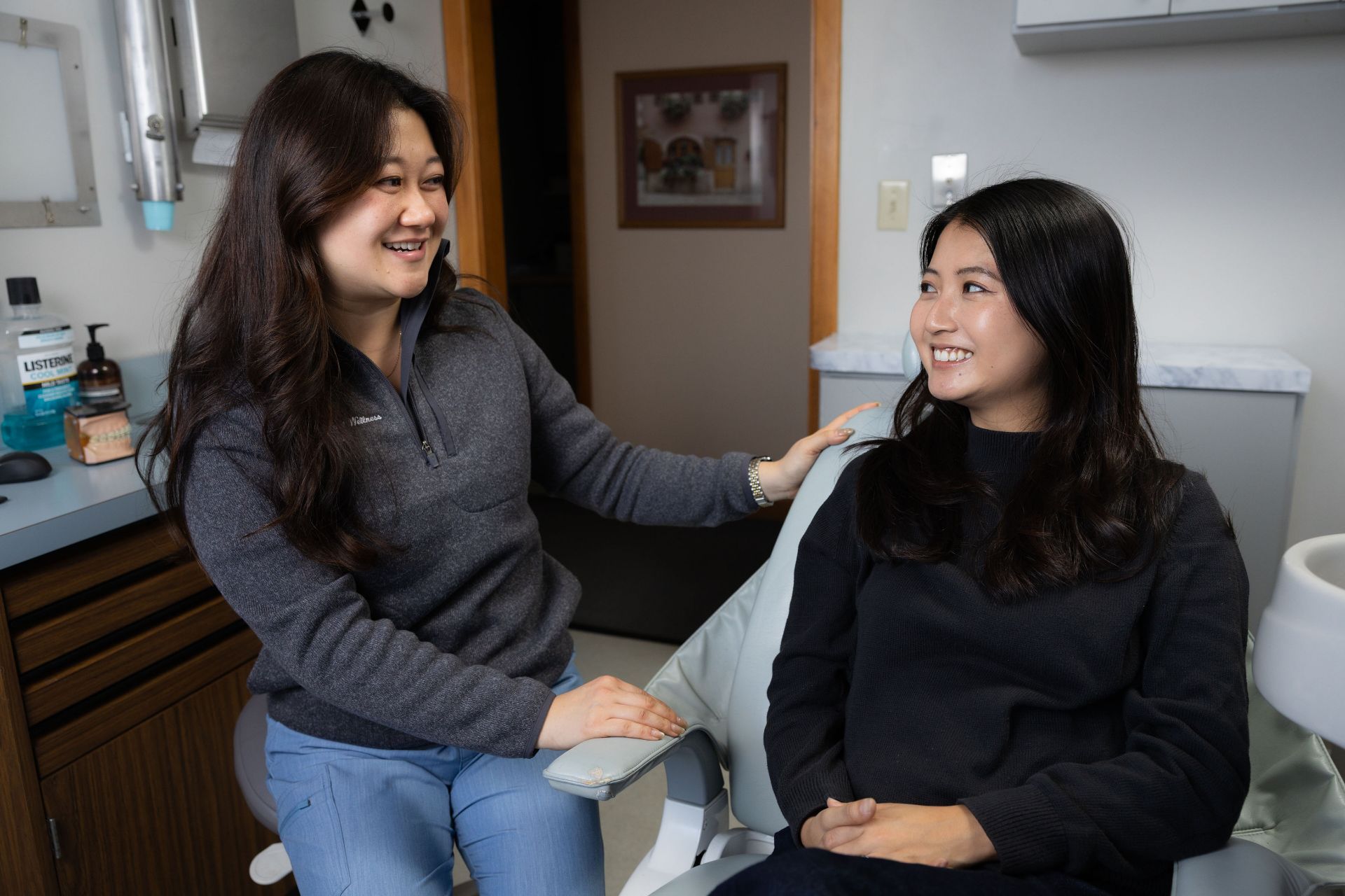 A dental professional in gray and blue scrubs smiling and talking with a female patient seated in a dental chair.