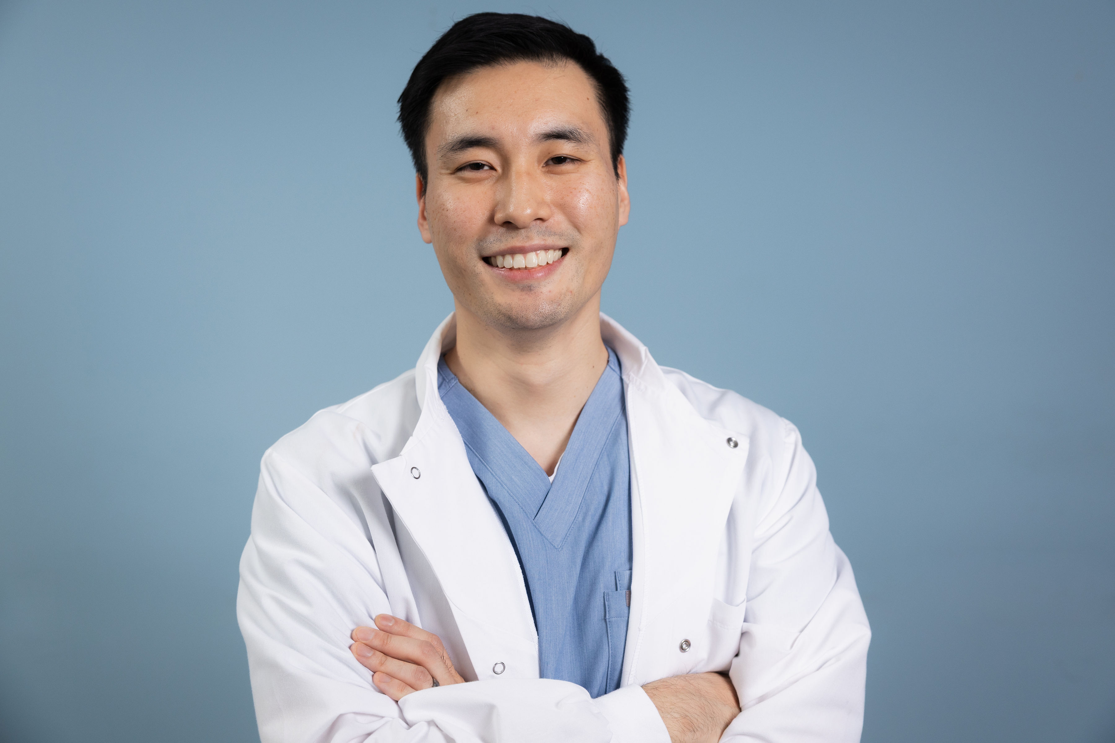 Smiling male doctor wearing blue scrubs and a white lab coat with arms crossed against a light blue background.
