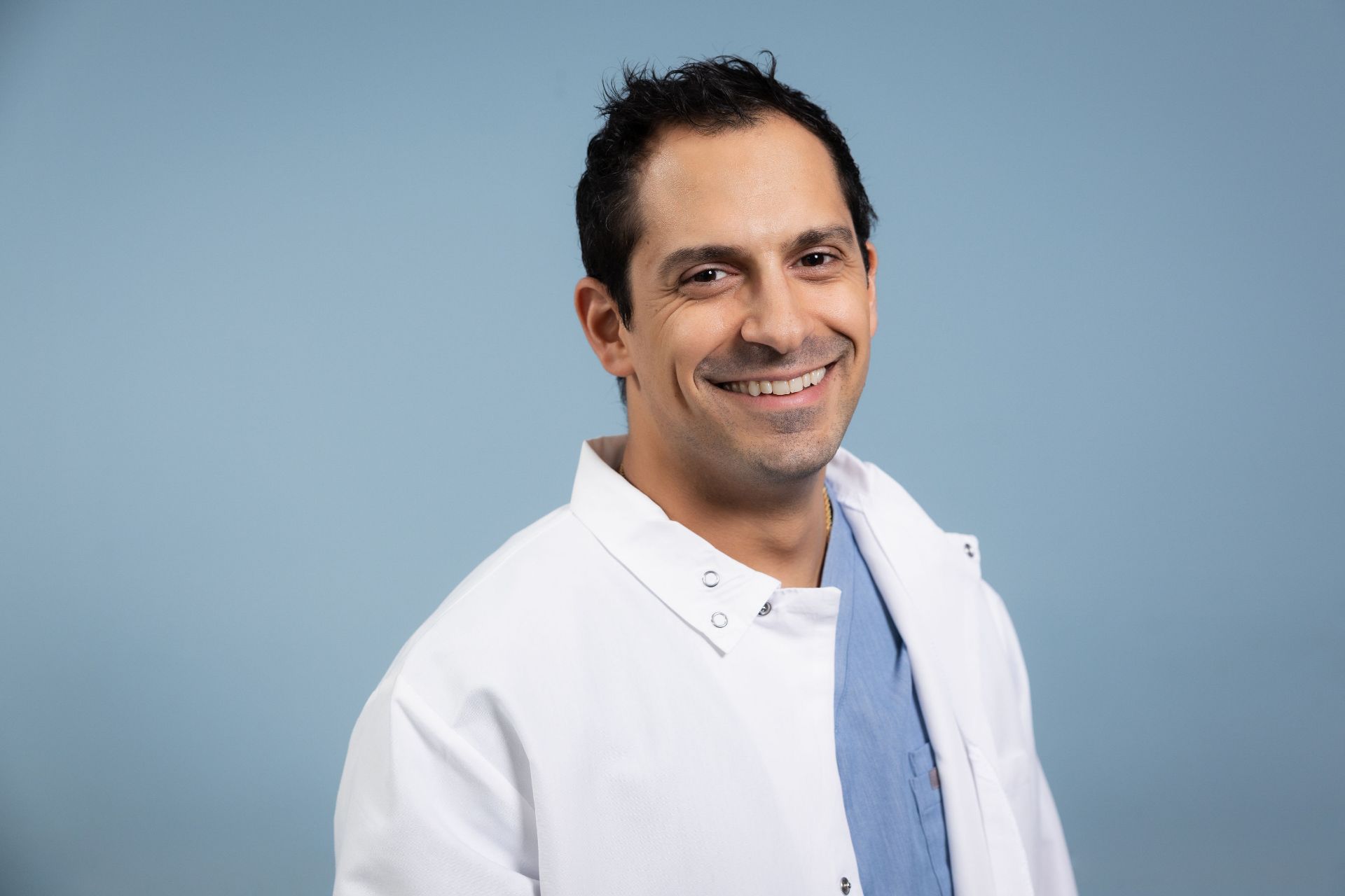 Smiling male doctor wearing white coat and blue scrubs against a light blue background.
