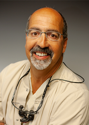 Smiling middle-aged male dentist wearing glasses and dental loupes around his neck.