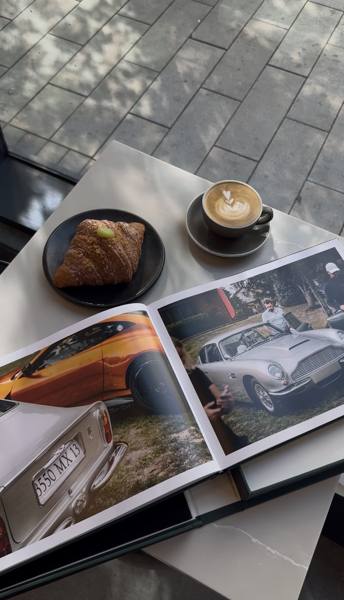 Open book showing photos of classic cars on a white table next to a croissant on a black plate and a cup of latte with heart-shaped latte art.