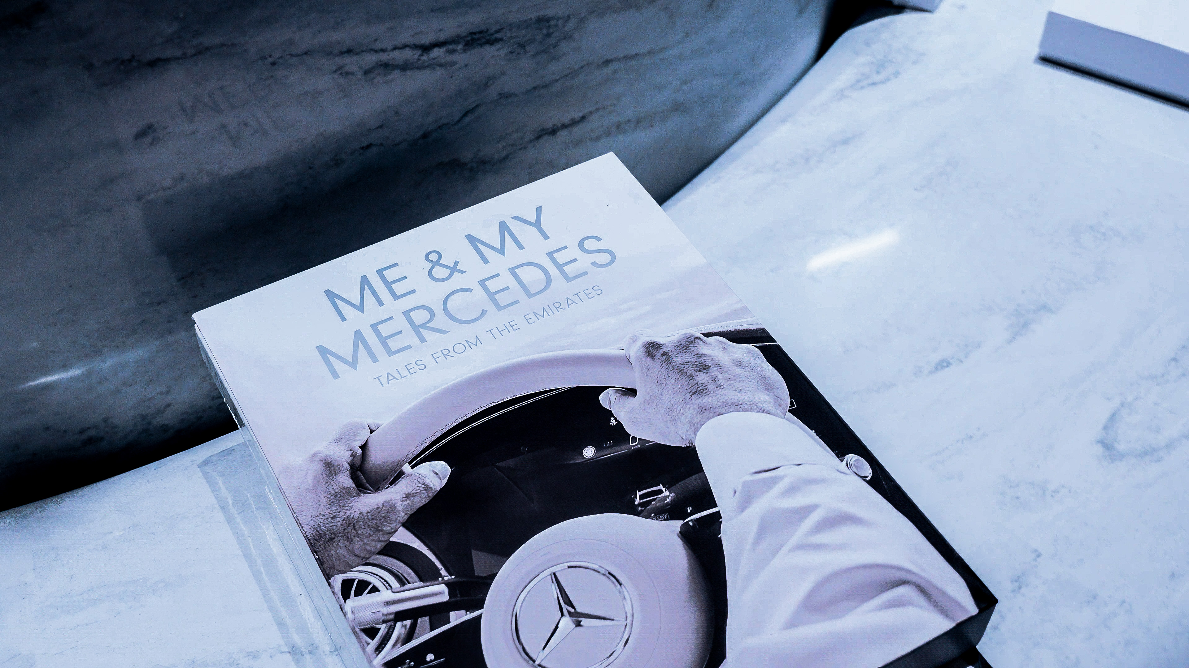 Book titled 'Me & My Mercedes: Tales from the Emirates' showing hands on a Mercedes steering wheel.