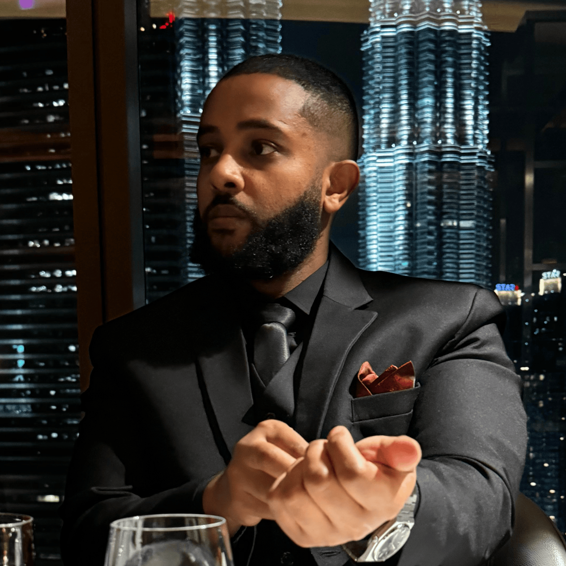 Man in a black suit adjusting his cufflinks with city lights and a tall illuminated building in the background.