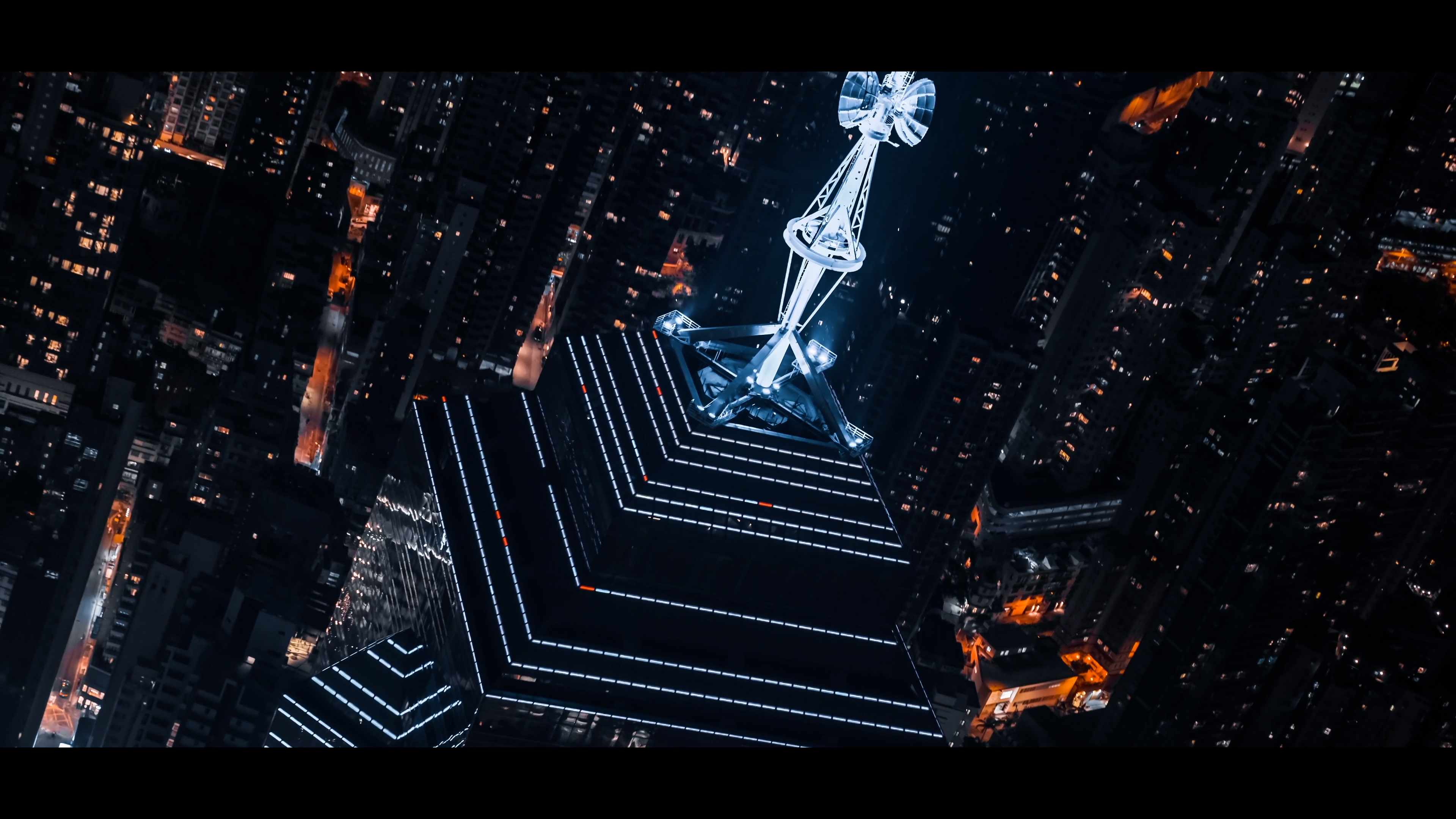 Aerial night view of a city skyscraper with illuminated lines and a bright antenna on top surrounded by city lights.