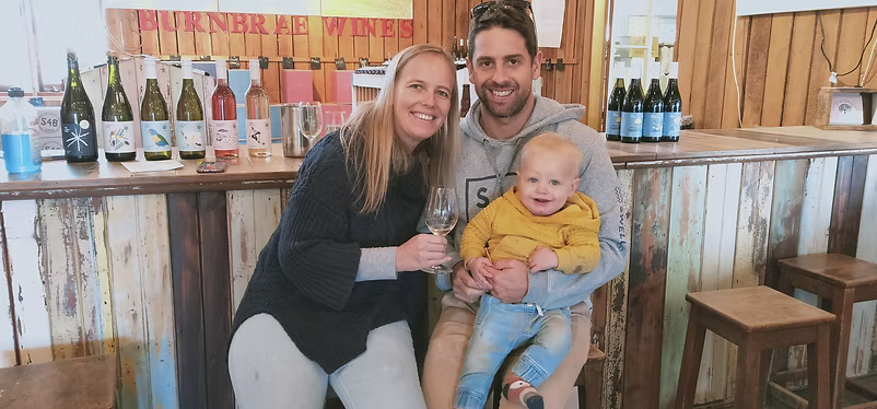 A smiling couple with a baby sitting on a wooden bar counter with various wine bottles displayed behind them.