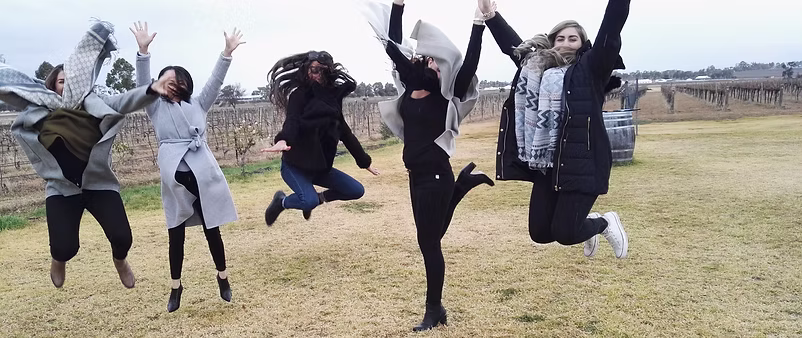 Group of five women outdoors on grass, jumping joyfully with arms raised, dressed in coats and scarves on a cloudy day.