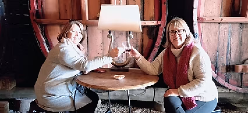 Two women sitting at a round wooden table clinking glasses in a cozy, rustic room with wooden barrels.