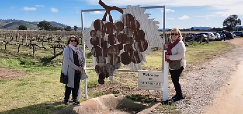 Two women standing on either side of a large metal grape cluster sculpture with a sign saying 'Welcome to Hahndorf' in a vineyard setting.