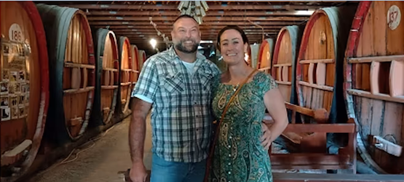 Man and woman smiling and posing together in a cellar with large wooden barrels lining both sides.