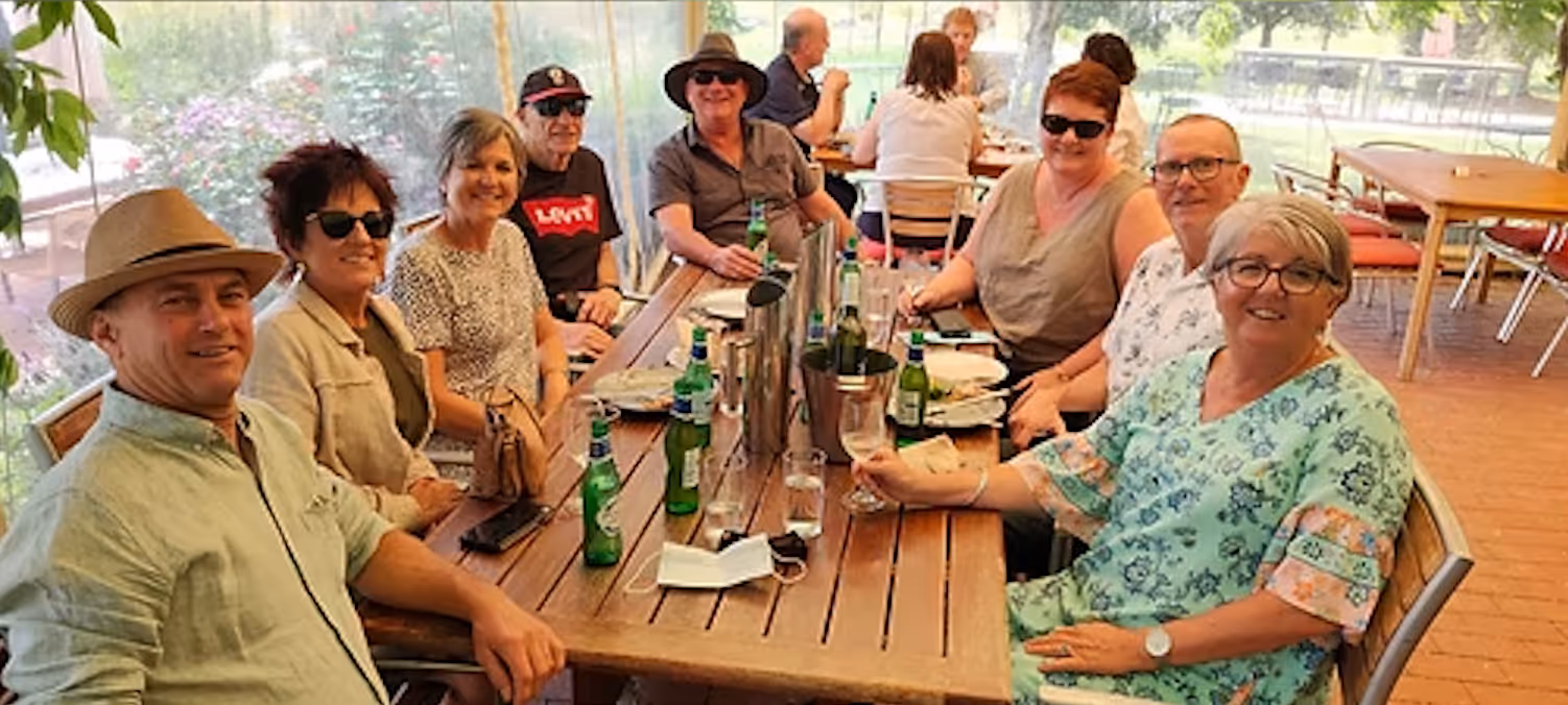 Group of eight adults sitting around a wooden table outdoors, enjoying drinks and smiling at the camera.