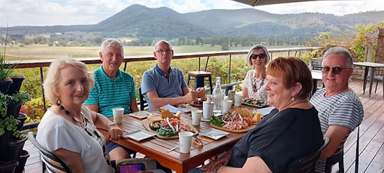 Group of six adults sitting around a wooden table outdoors with mountains in the background, enjoying food and drinks.