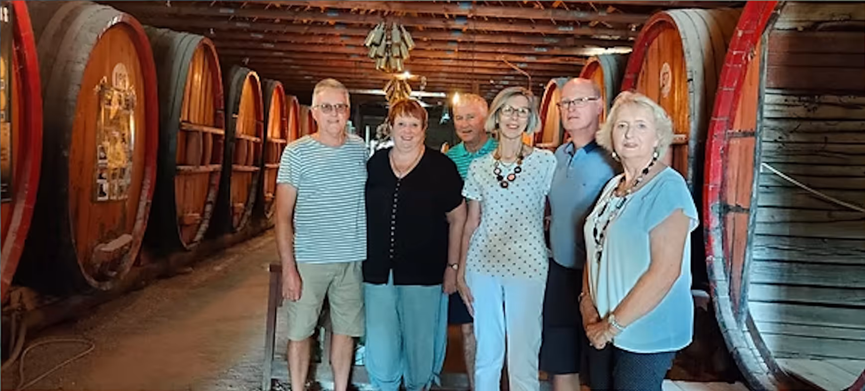 Six people standing inside a wine cellar with large wooden barrels lining both sides.