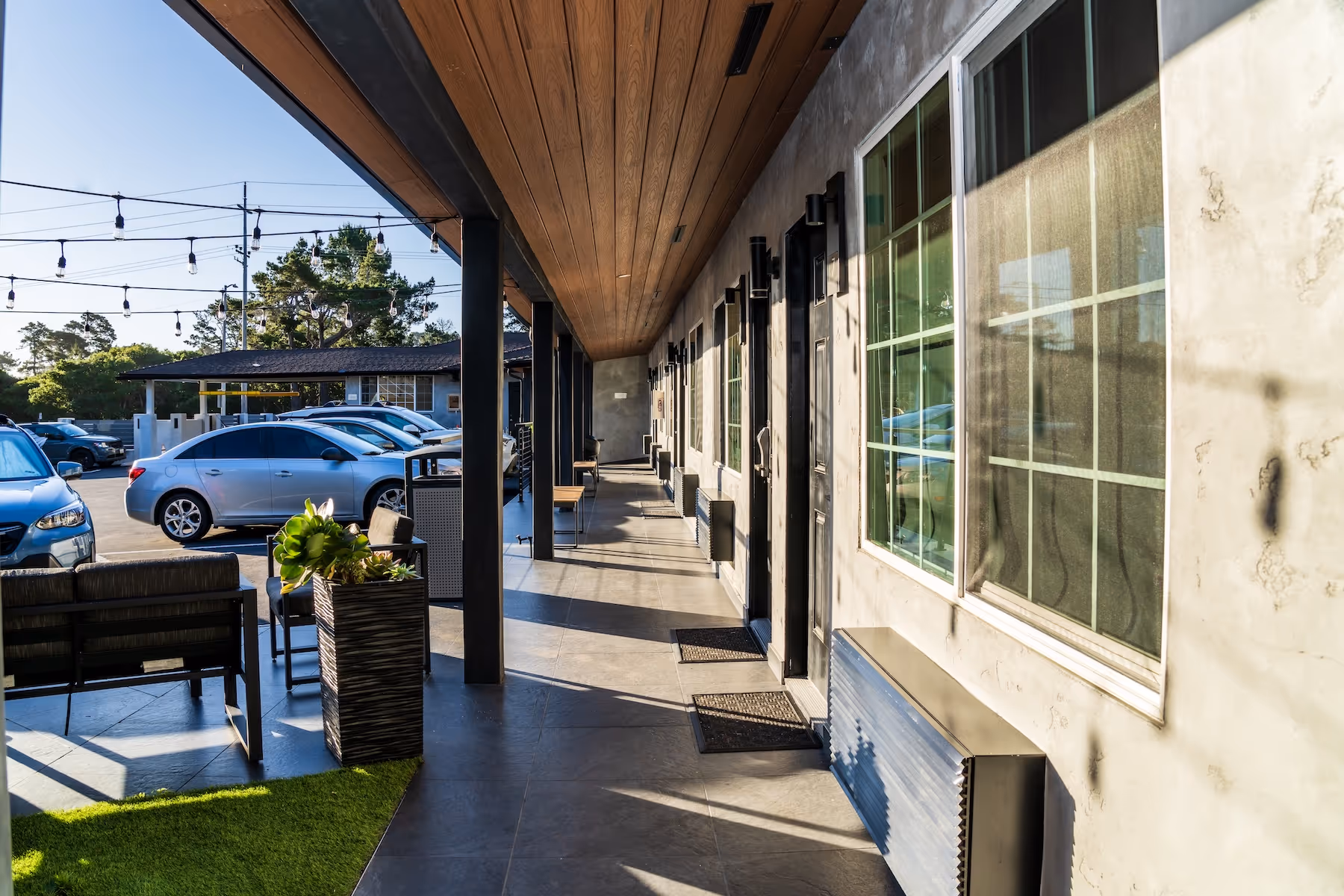 Outdoor motel corridor with black doors, large windows, and potted plants, next to a parking lot with cars under string lights during daytime.