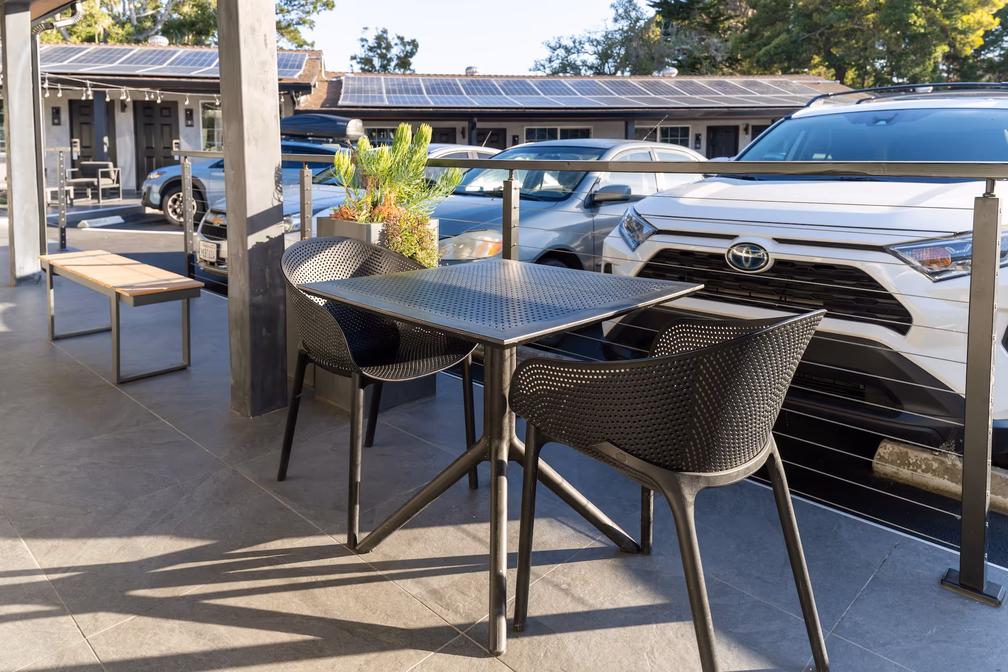 Outdoor patio area with a black metal table and two perforated black chairs, overlooking parked cars including a white Toyota SUV.