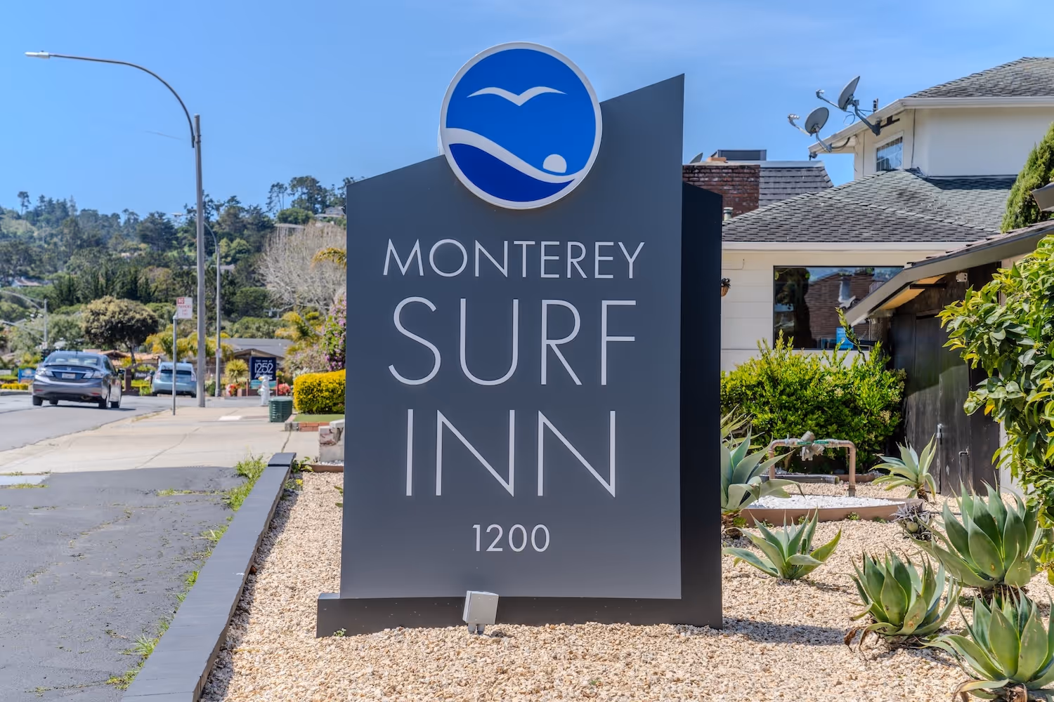 Monterey Surf Inn sign with a wave and bird logo, surrounded by gravel and desert plants near a street.