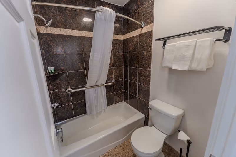 Clean bathroom with a white bathtub, shower with white curtain, dark tiled walls, toilet, and shelves with folded towels.