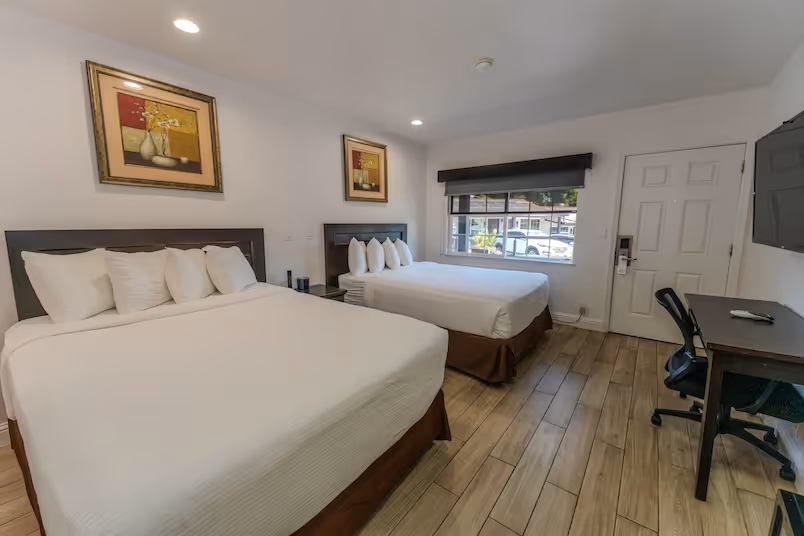 Hotel room with two queen beds, white linens, a desk with a chair, a wall-mounted TV, and a window with a view of parked cars outside.