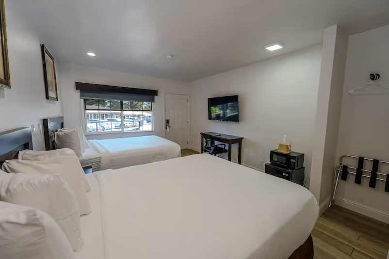 Bright hotel room with two double beds, a window showing parked cars, a wall-mounted TV, a small desk, and a microwave on a stand.