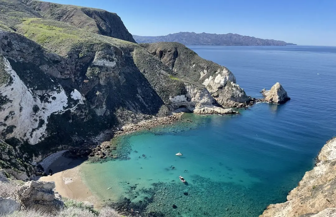 Clear blue cove with rocky cliffs and a small sandy beach under a bright blue sky.